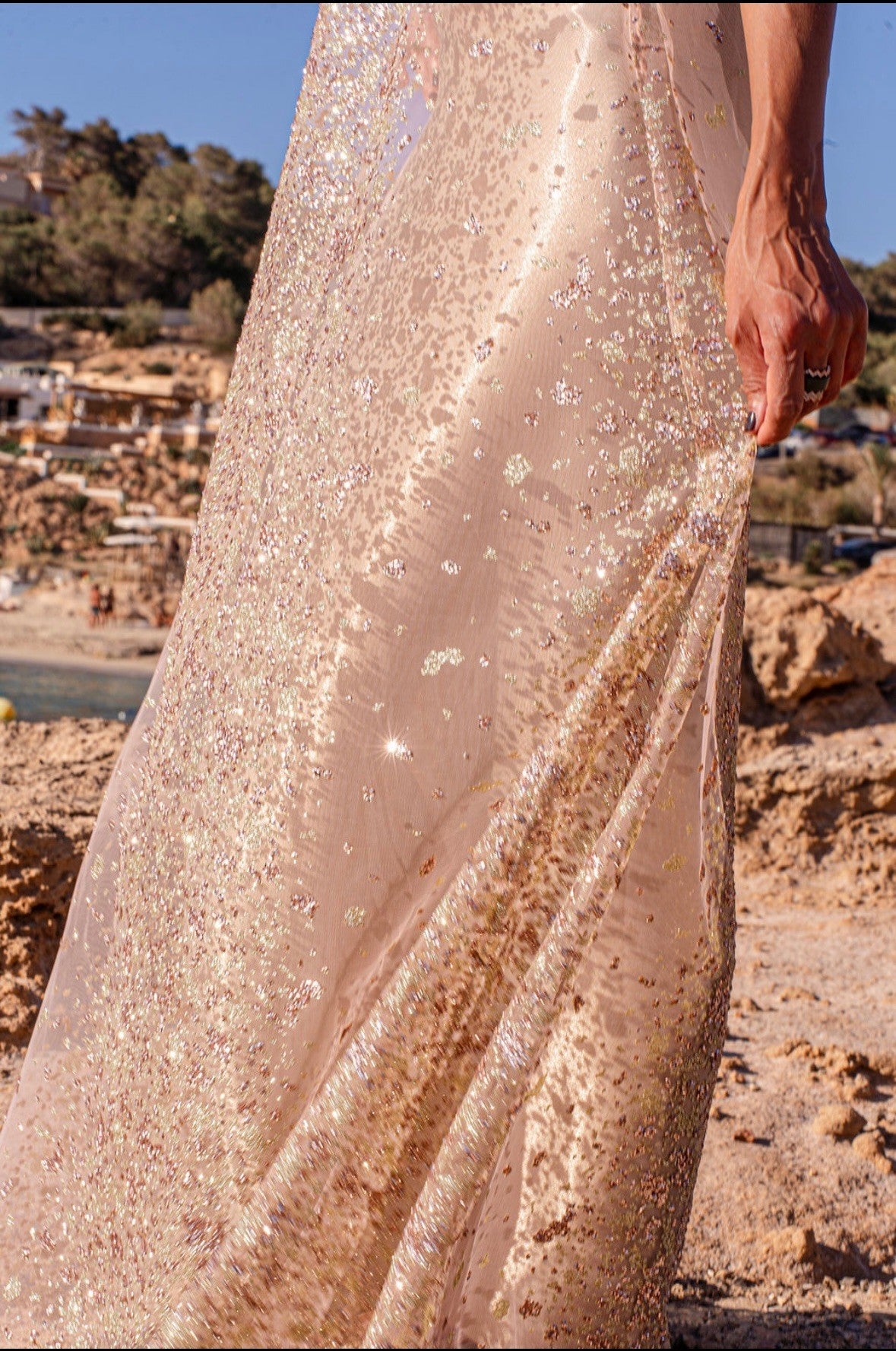 A close-up of the bottom of the sheer pink dress with pink, gold and silver glitter embellishments, worn over an oyster coloured slip, next to the beach