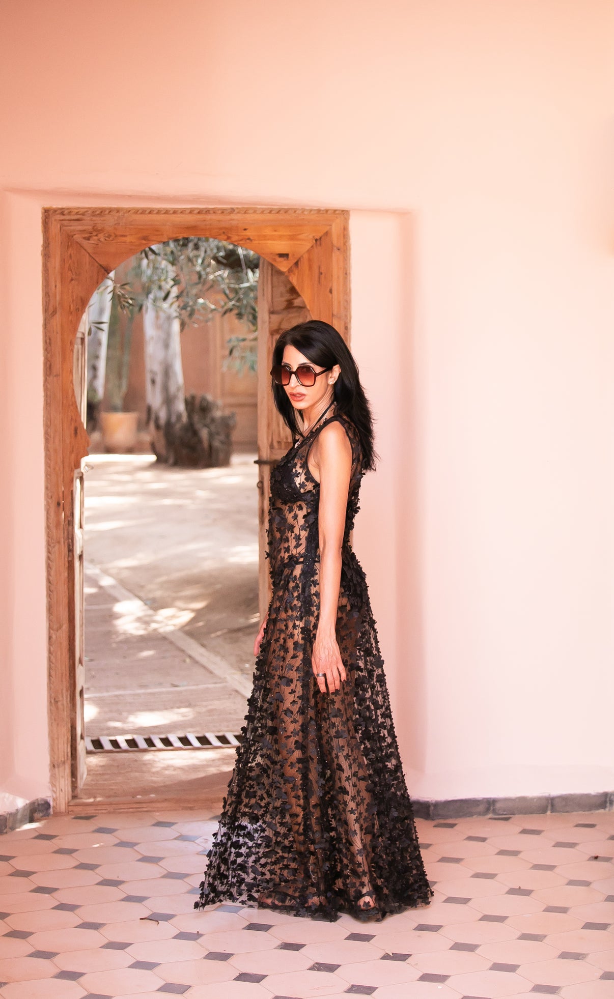 Full-length view of the Daisy Noir black sheer gown with floral appliqué, styled with oversized sunglasses in a Moroccan-inspired setting