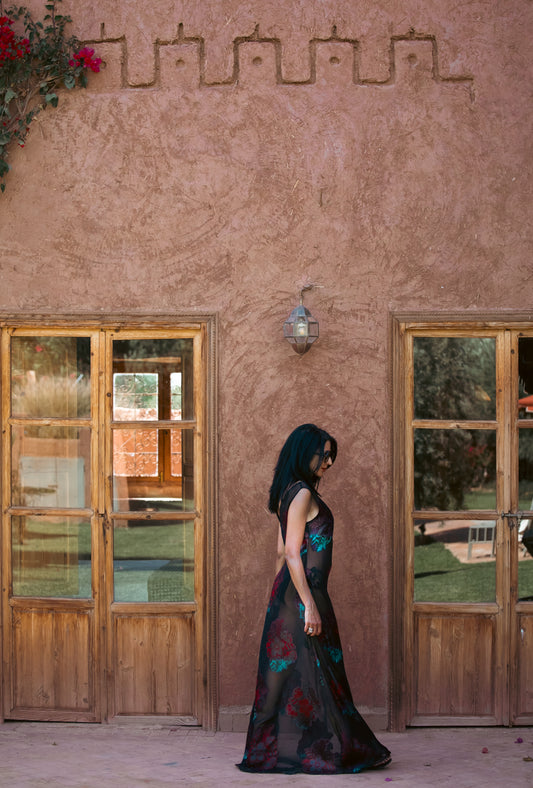 Side view of the Dusky Rose sheer black gown with red and teal floral embroidery, photographed against an earthy Moroccan wall