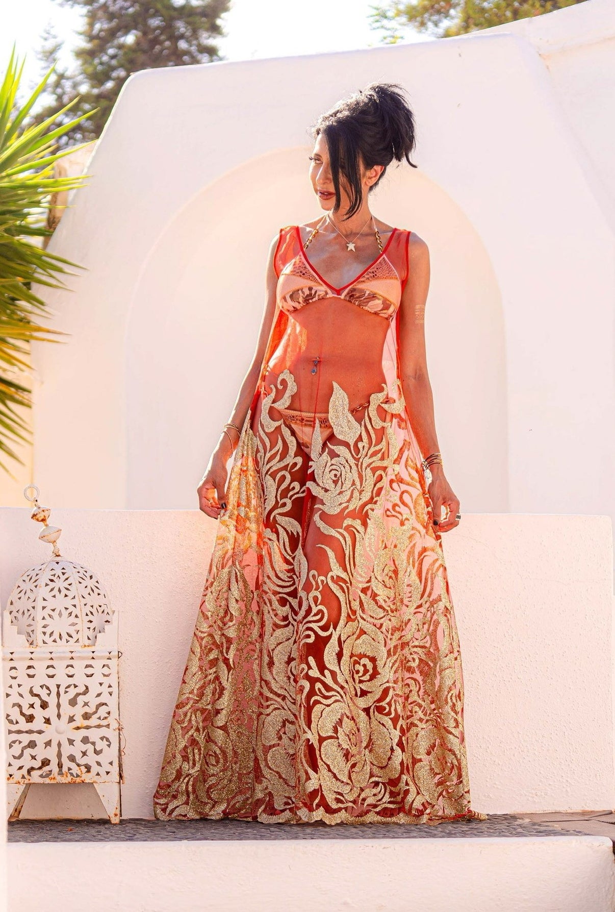 A woman standing against a white stone background wearing the Enchanted Ember dress by Preema Aura - a sheer red dress with gold glitter detailing