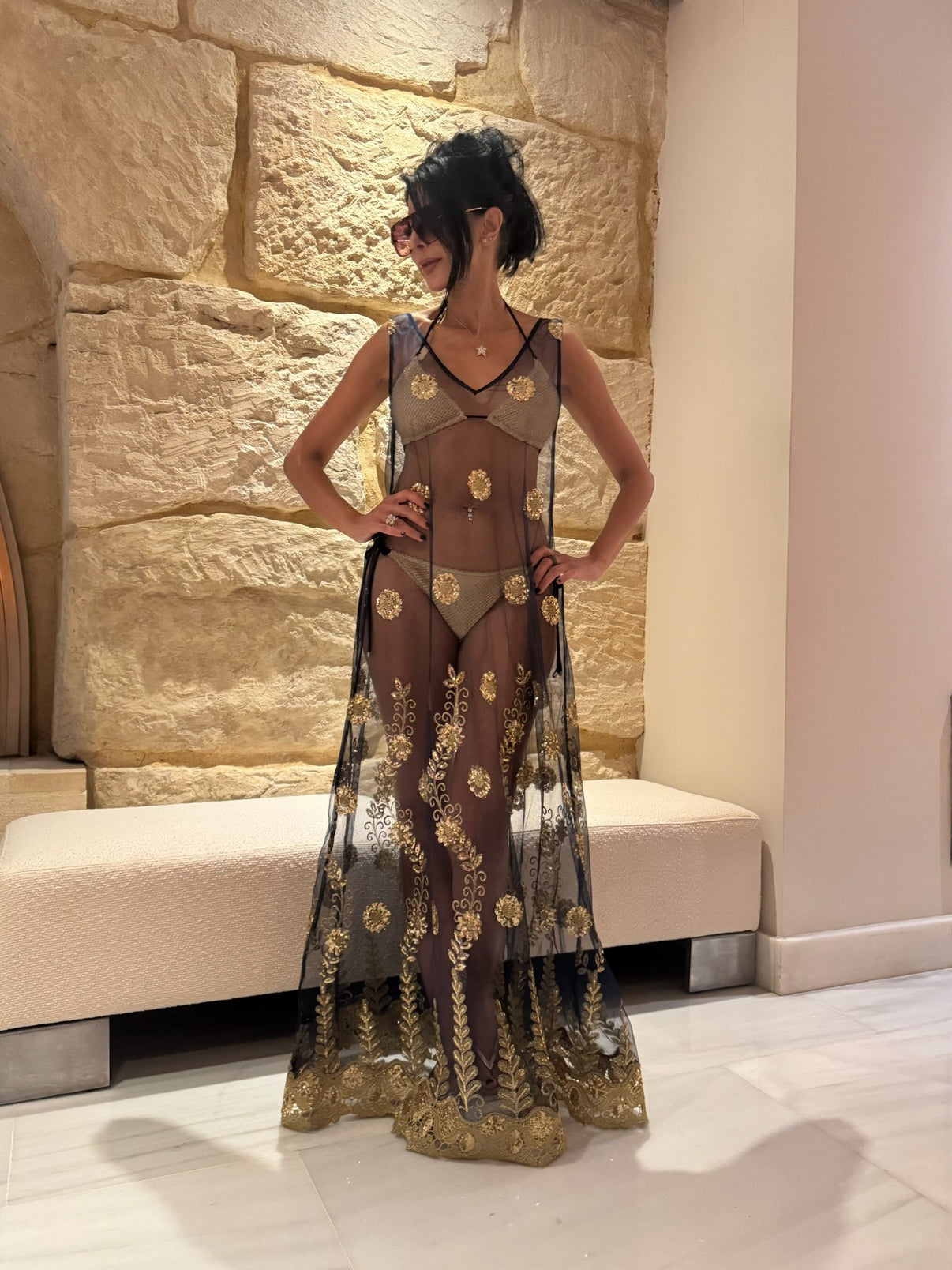 A person wearing a navy sheer gown with golden embellishments standing in a room with stone walls.