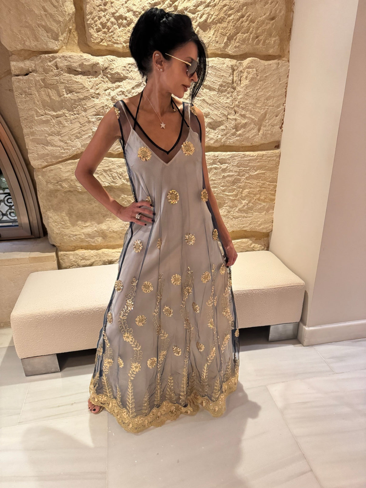 A woman wearing a cream slip under a sheer navy maxi dress with gold embellishments, standing in front of a stone wall