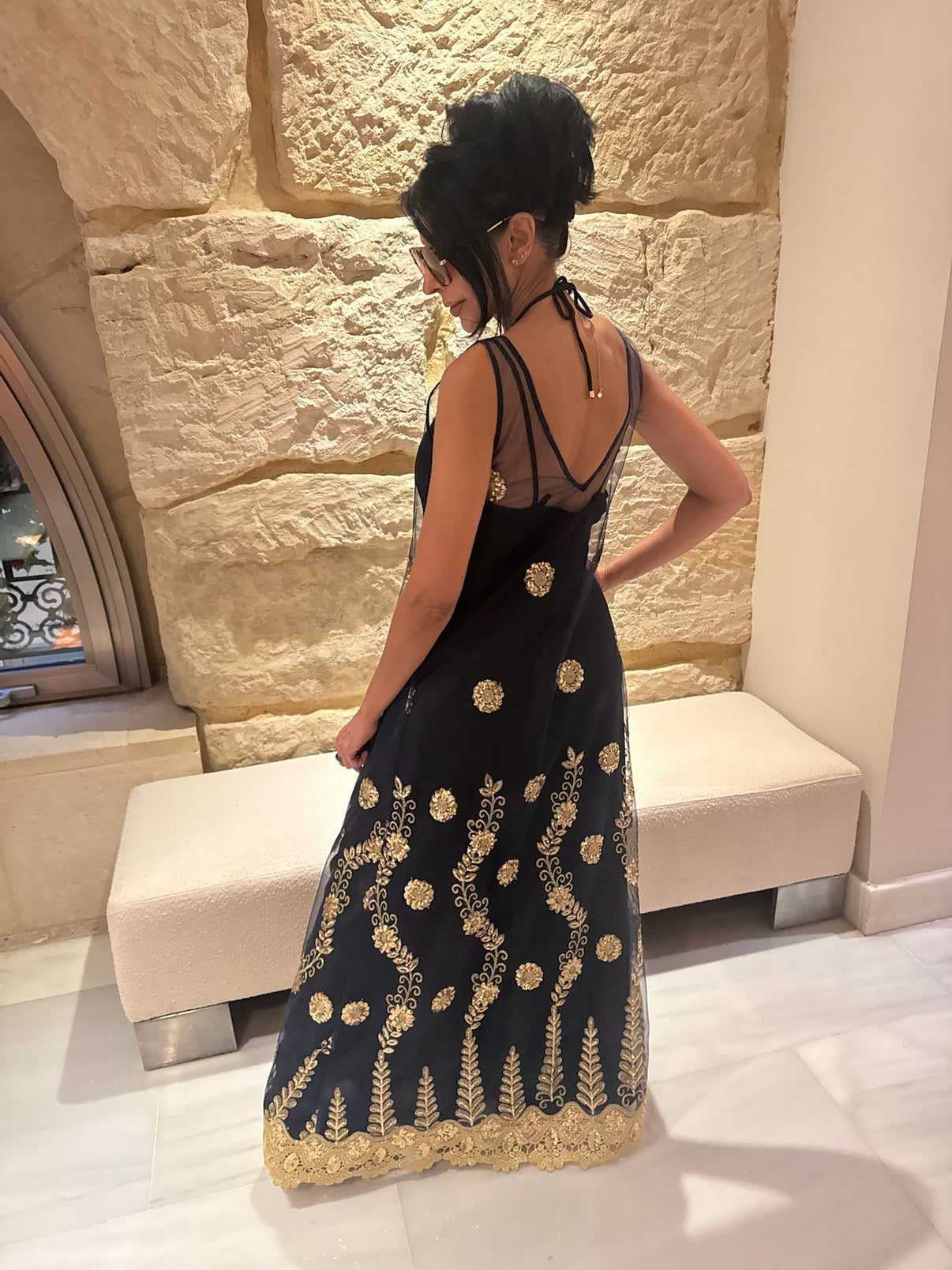 A rear view of a woman wearing a sheer navy dress with gold embellishments, standing in front of a stone wall
