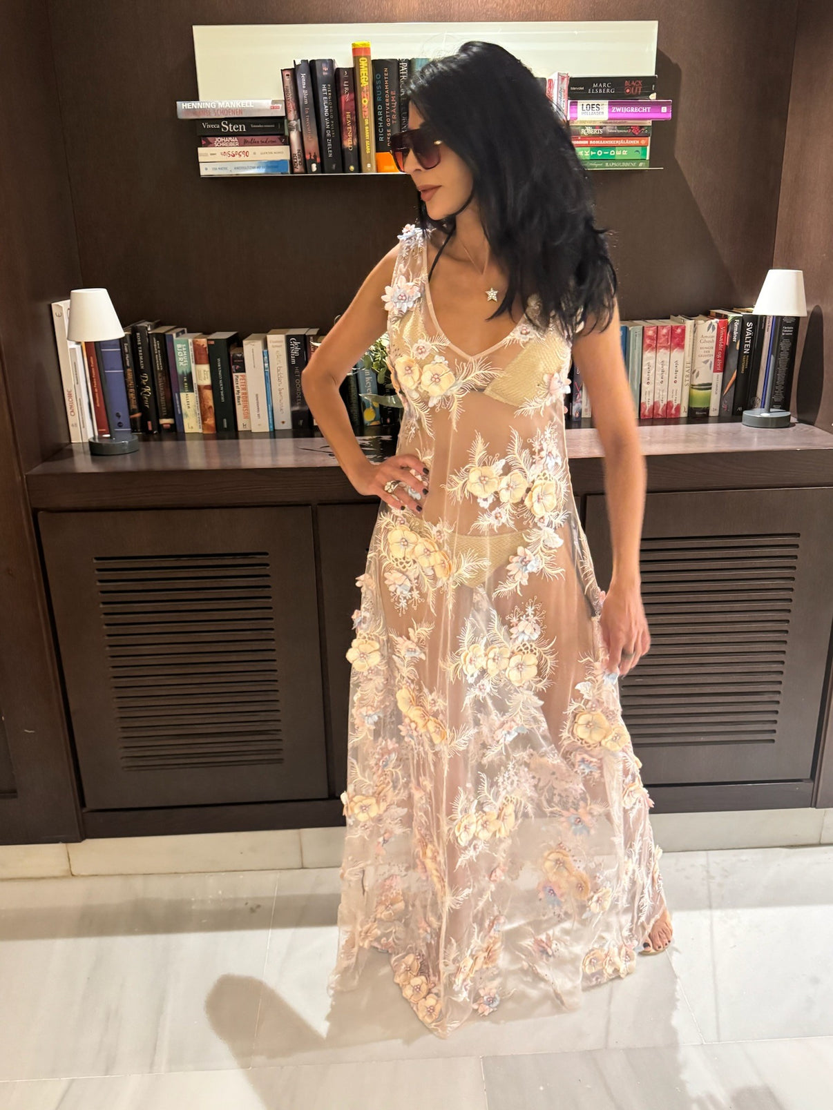 A woman standing in a room with an array of books, wearing a sheer, floral tulle dress with pale pink and cream colors, paired with sunglasses and a hand on her hip.