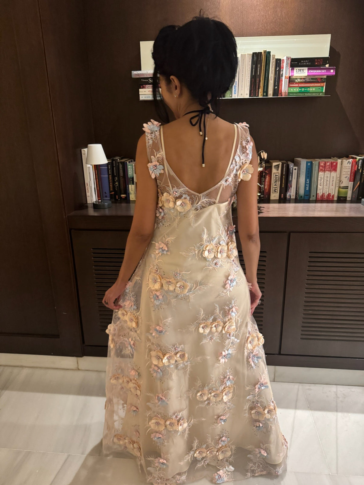 A woman wearing a textured, sheer maxi dress with pink, cream and blue floral embellishments, over a cream slip. She is facing away from the camera, towards a bookshelf.