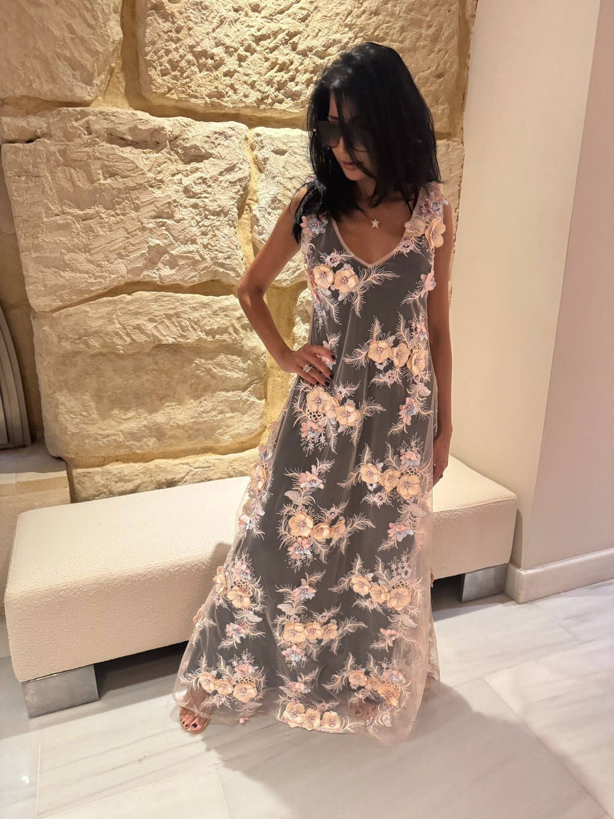 A woman wearing a sheer pink maxi dress with cream, pale pink and blue floral embellishments, over a black slip, whilst standing in front of a stone wall