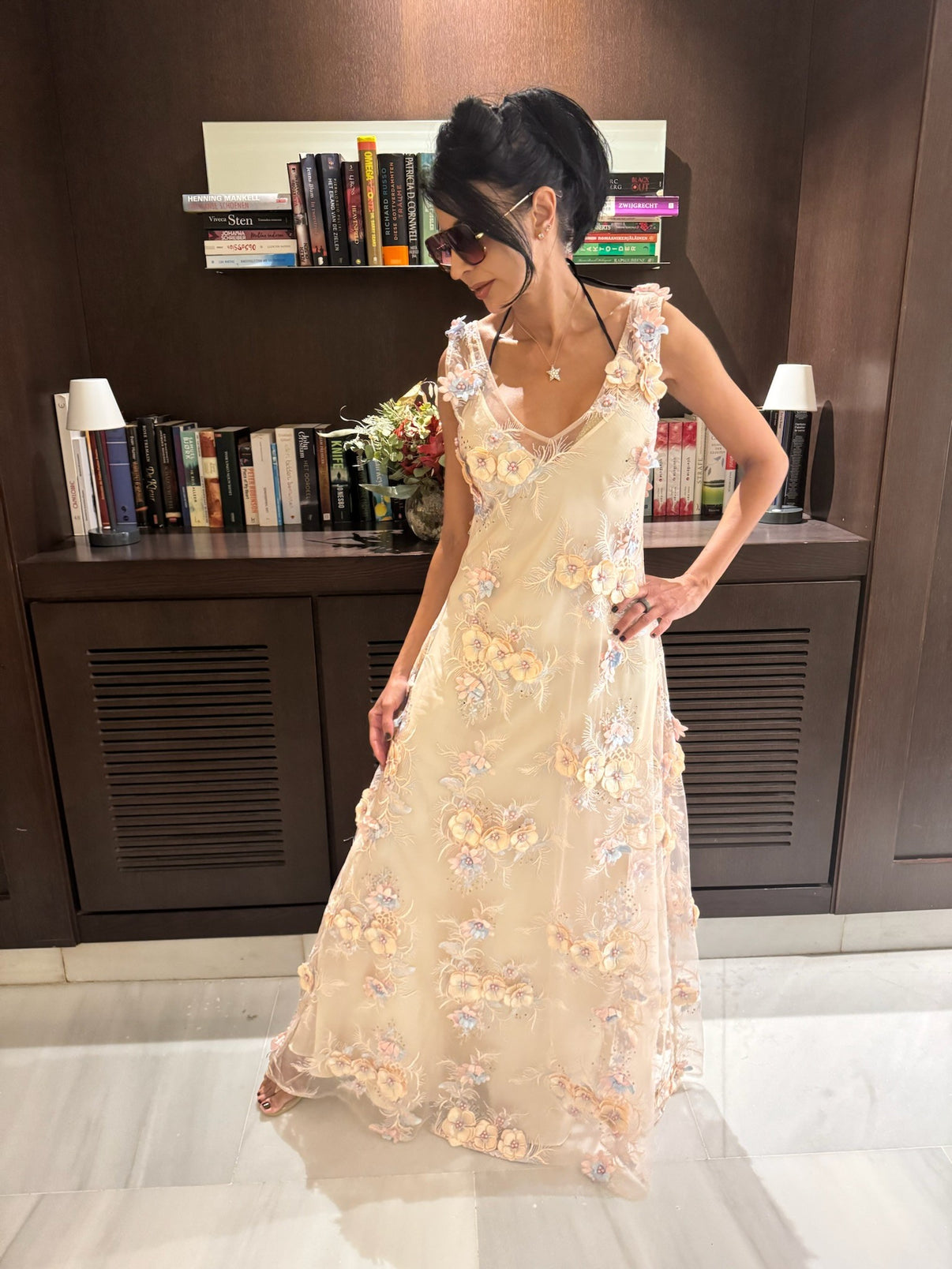 A woman wearing a textured sheer maxi dress with floral embellishments, over a cream maxi dress whilst standing in front of a bookshelf.