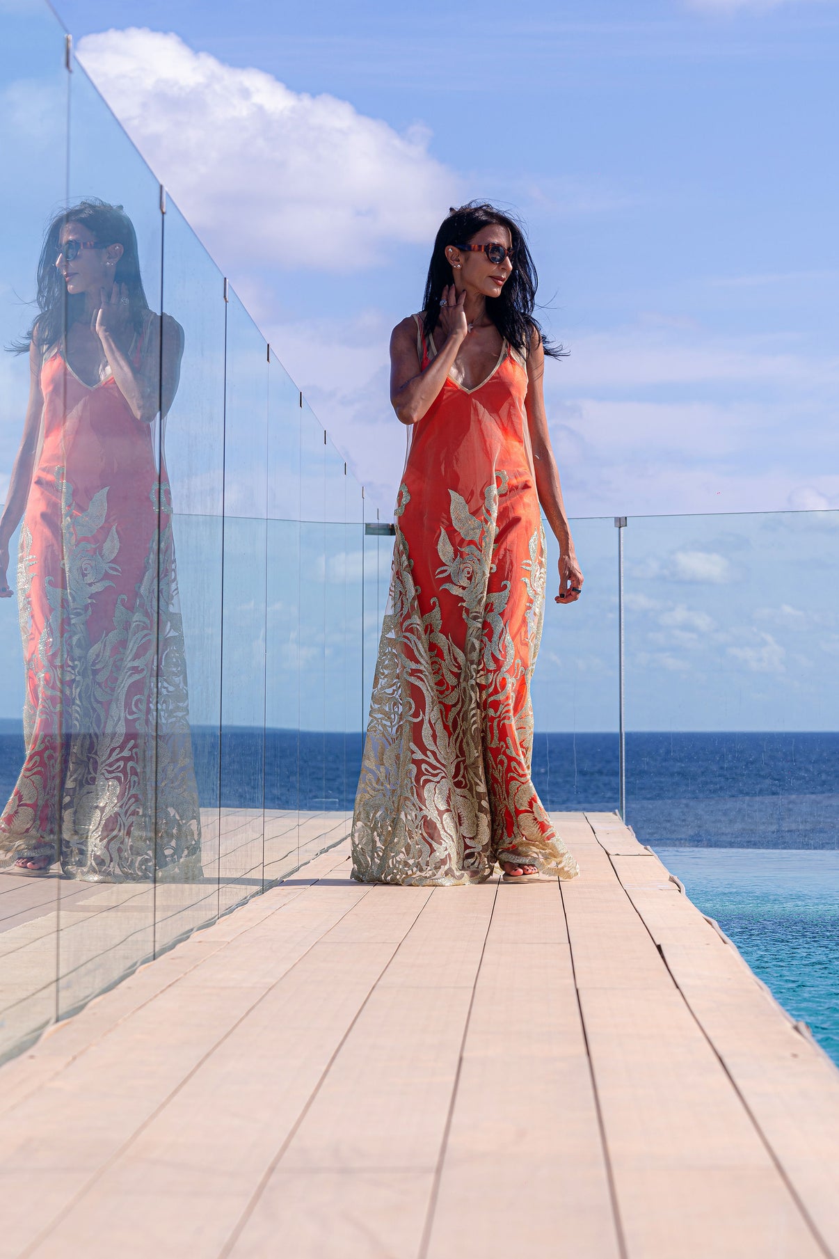 Model strolling along the side of a swimming pool, with the sea in the background, in the Citrine Essence dress with a Red Satin Slip, capturing relaxed coastal elegance.
