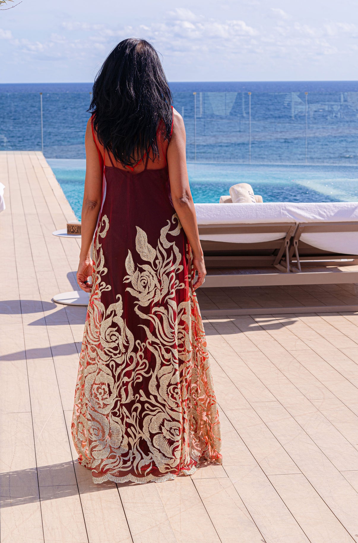 Back view of the Enchanted Ember dress with a black satin slip underneath, as the model towards a swimming pool by the sea.