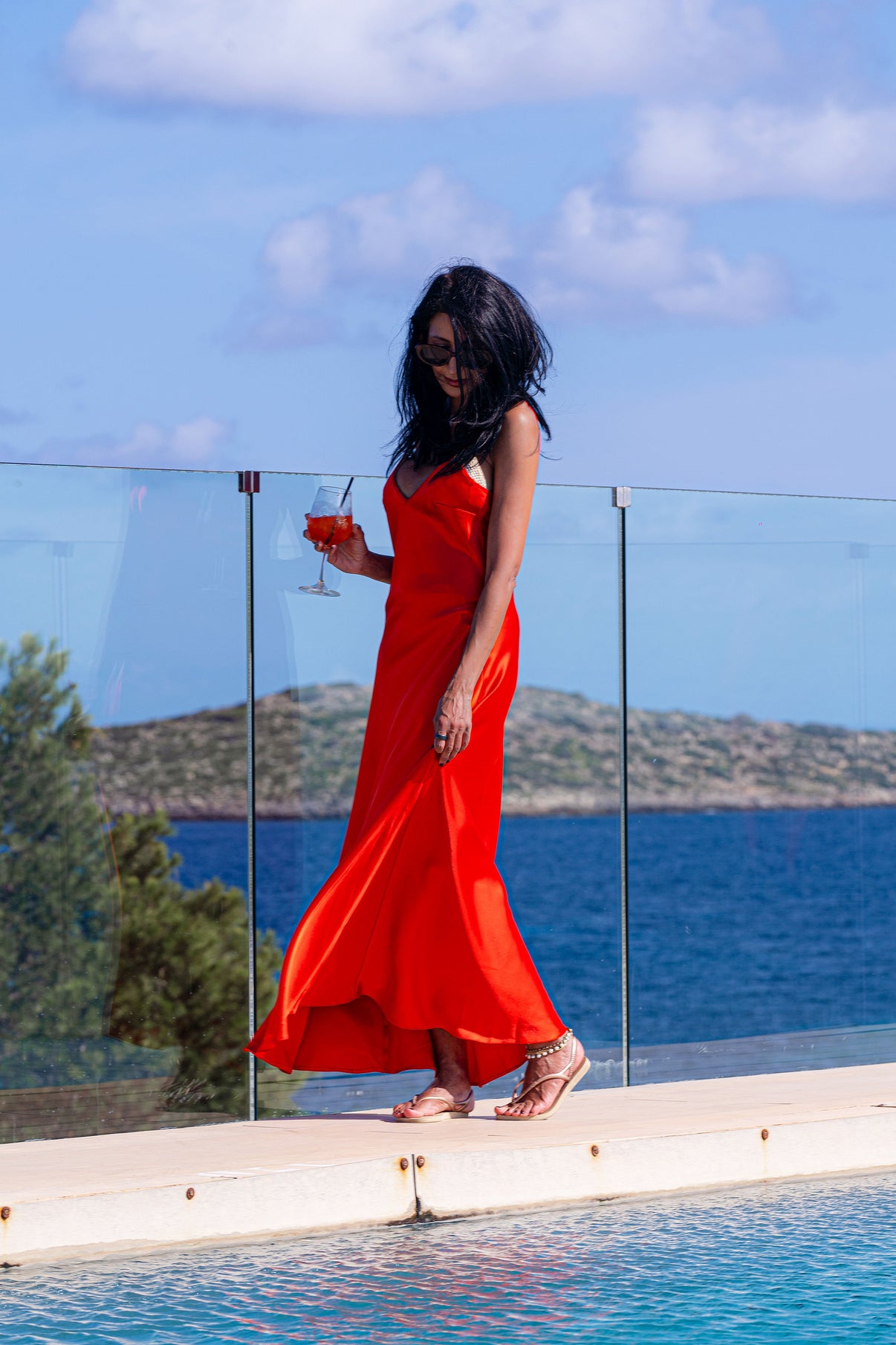 A woman in a red satin slip dress standing by a pool with a glass of wine, wearing heels and with her hair down.
