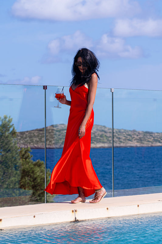A woman in a red satin slip dress standing by a pool with a glass of wine, wearing heels and with her hair down.