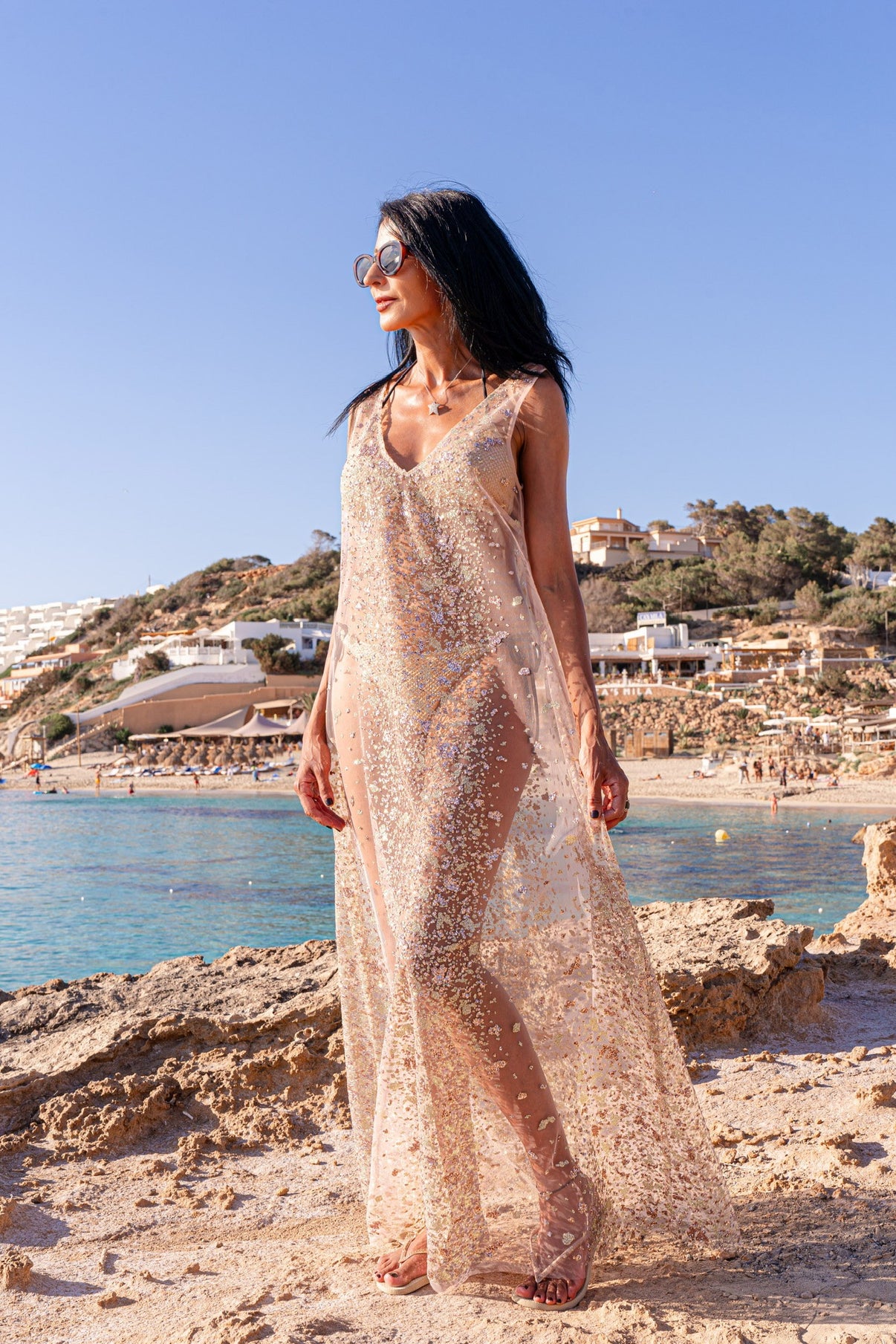 A woman standing on a rocky beach wearing a pink, sequin, maxi dress with a V-neckline and sleeveless top.