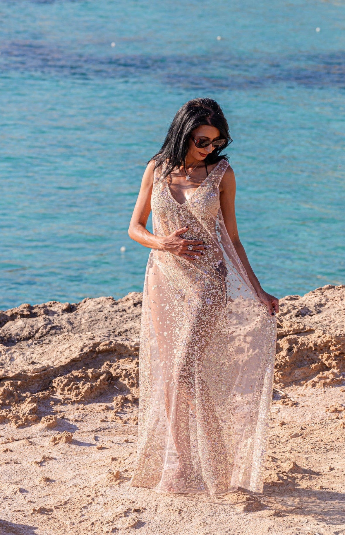 A woman wearing a sheer pink maxi dress with glitter embellishments, worn over a gold bikini, standing on some rocks in front of an azure blue sea