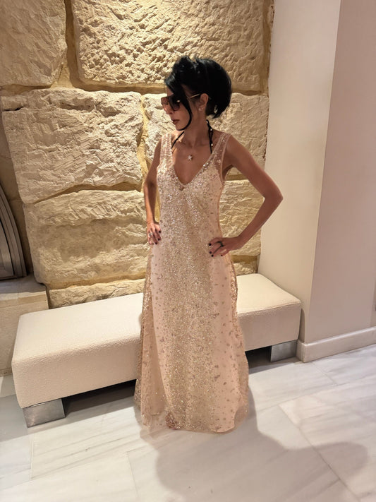 A woman wearing a cream slip under a sheer pink maxi dress with gold, pink and silver glitter embellishments, whilst standing in front of a stone wall
