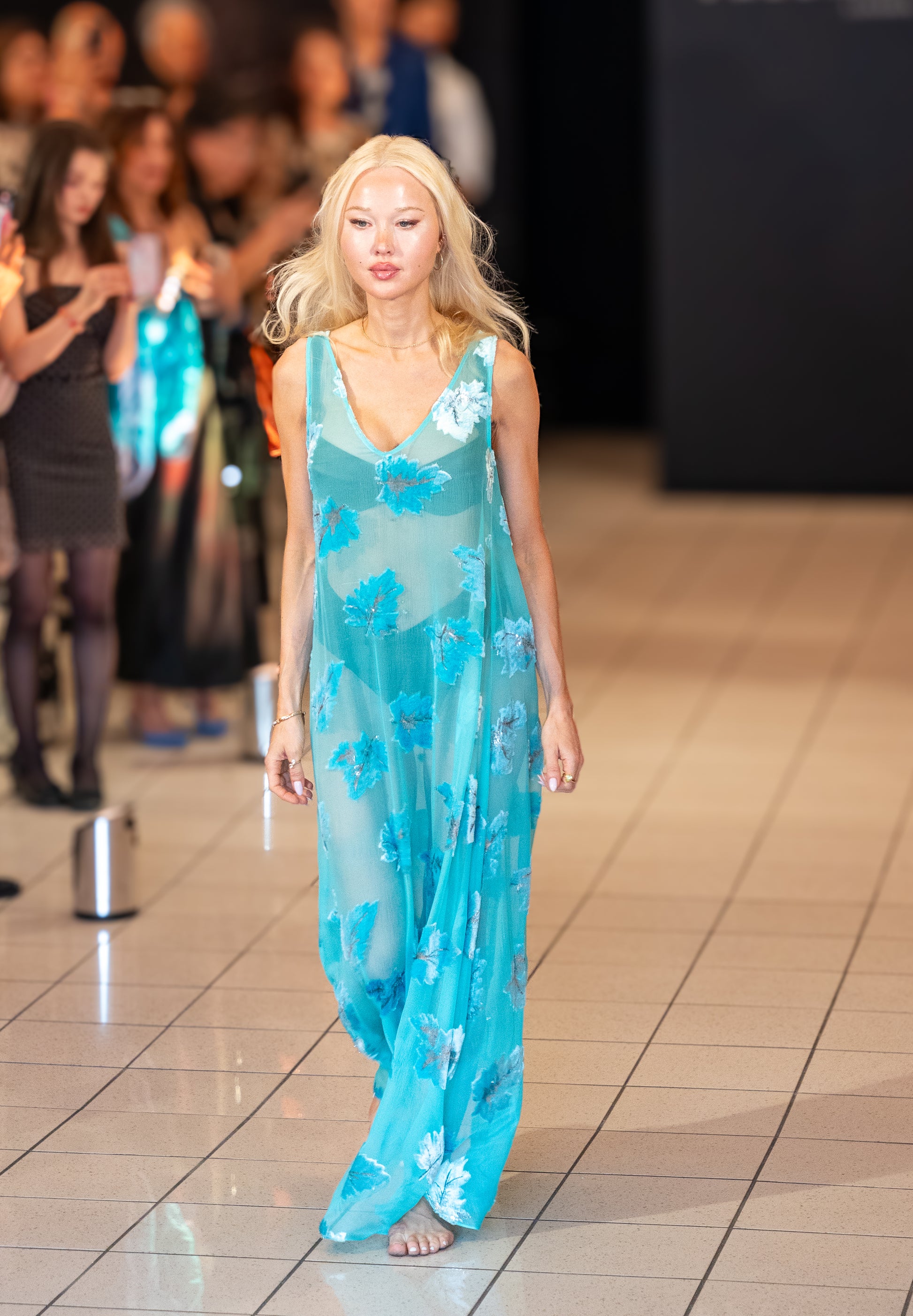 Model walking runway in Soul Cascade by Preema Aura, turquoise sheer silk maxi dress with soft floral appliqué, luxury London fashion.