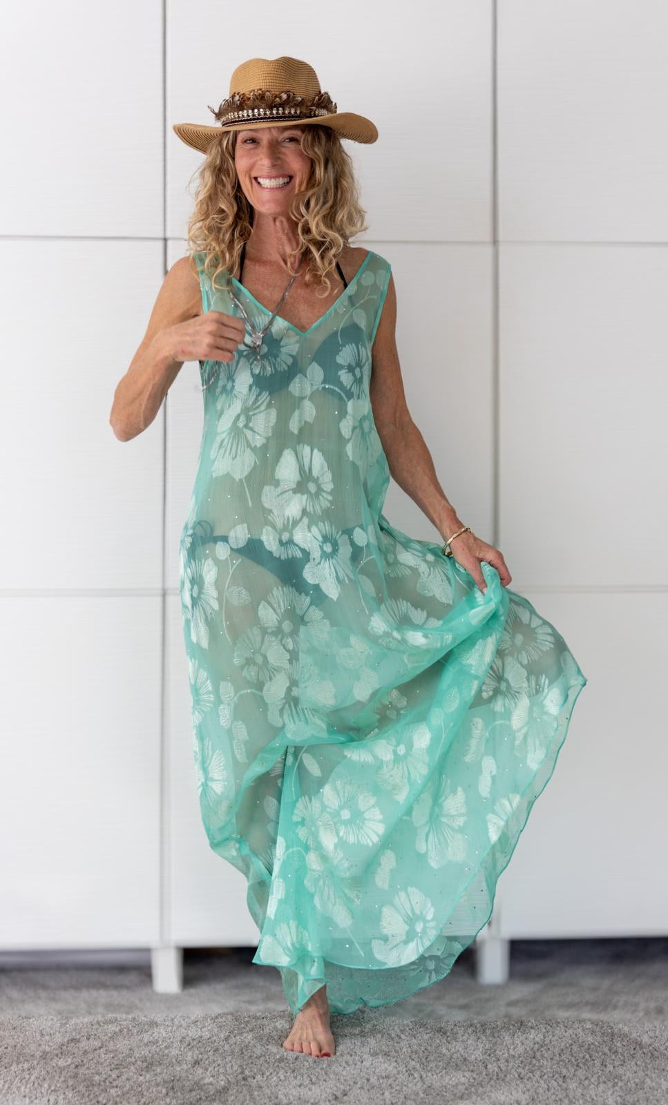 Woman wearing the Soul Eden dress by Preema Aura, a sheer turquoise one-off design from the Soul Series, styled with a straw hat to highlight its flowing fabric and large white floral print, embodying effortless resortwear elegance.