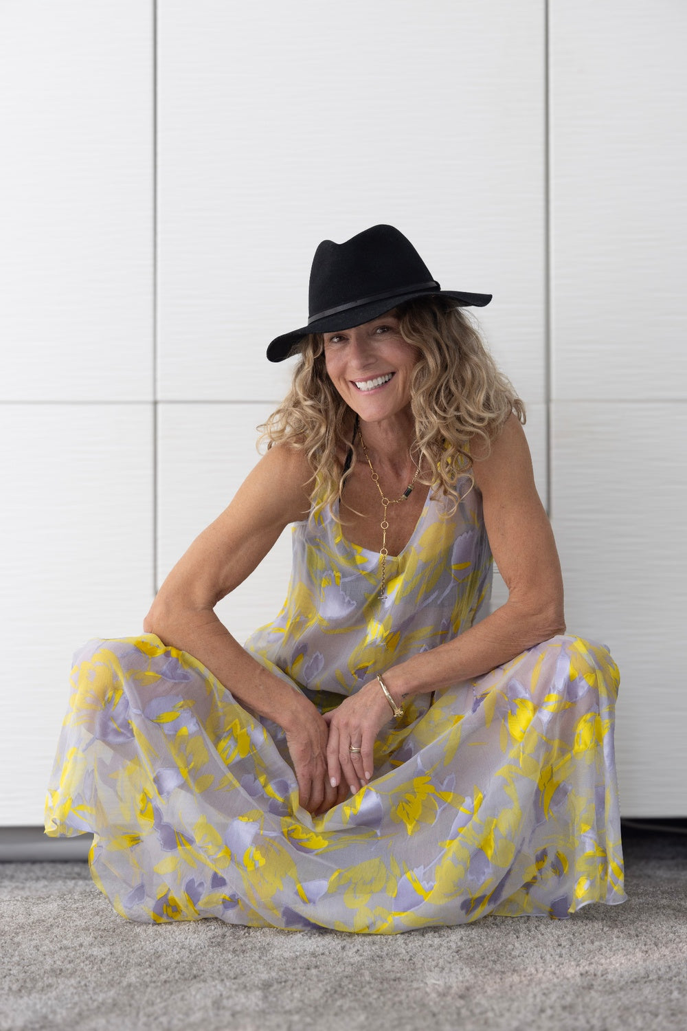 Preema Aura Soul Radiance dress modelled seated, showcasing the sheer lilac and yellow floral fabric and relaxed resortwear silhouette, styled with a black hat.