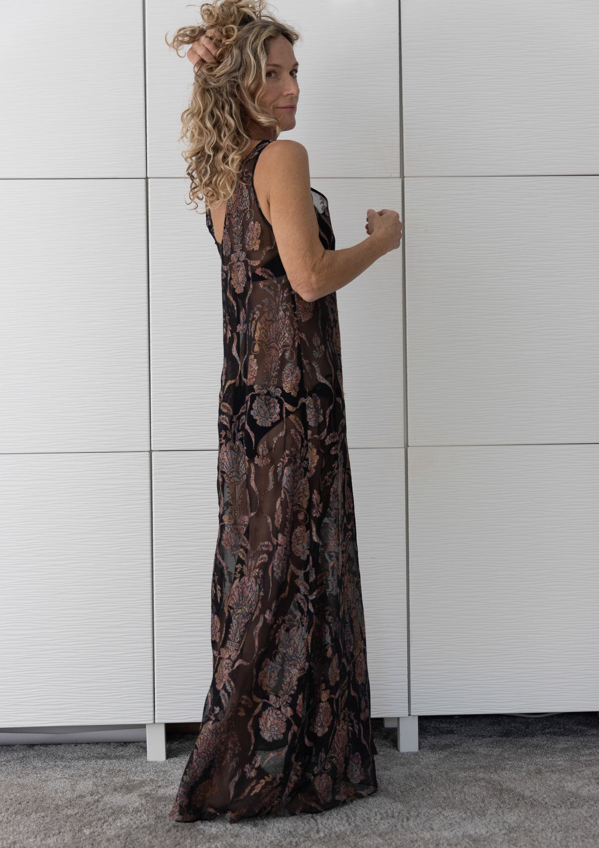 Side view of Soul Sway sheer black silk A-line slip dress layered over a bikini, showcasing its fluid drape and elegant paisley-style copper-bronze pattern in natural indoor light